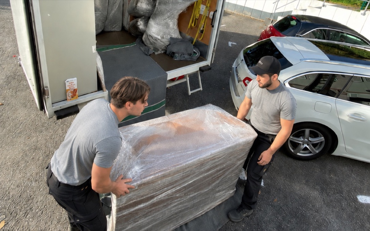 Umzug: zwei Mitarbeiter von Ceylan tragen ein in Folie verpacktes Möbelstück zum Transporter